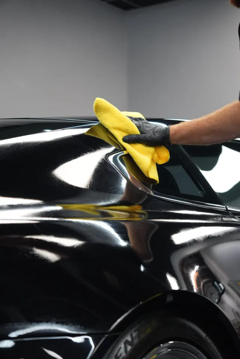 A black-gloved hand wipes the rear roof and quarter panel of a black car with a yellow microfiber cloth.