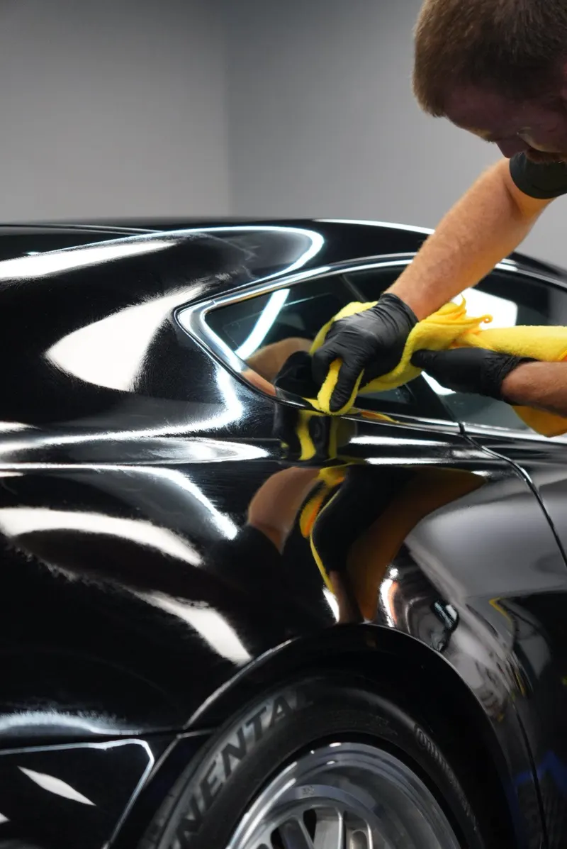 A person in gloves leans over to wipe the rear quarter panel of a black car with a yellow microfiber cloth, the wheel below.