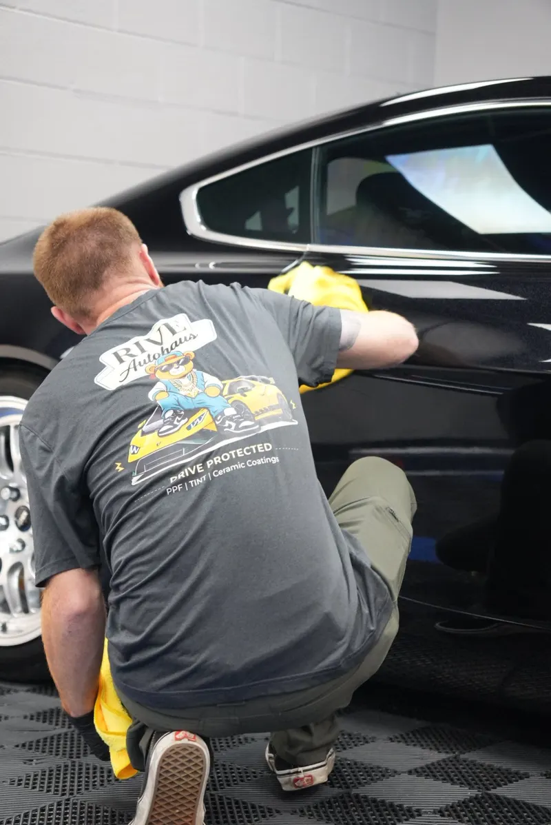 A technician in a Rive Autohaus shirt crouches beside a black car, wiping the door panel with a microfiber cloth in a shop bay.