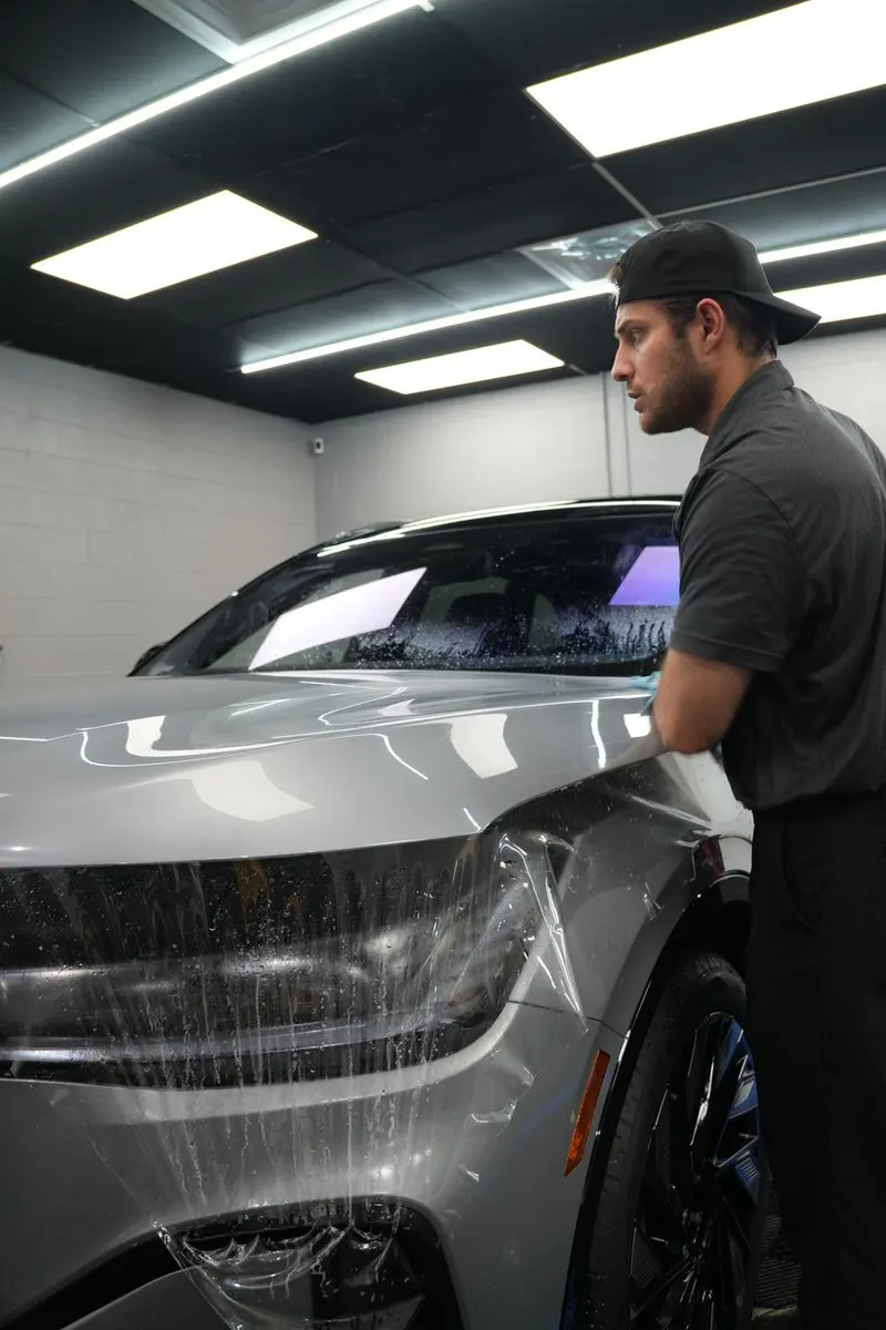 A technician smooths film against the side of a vehicle in a shop bay.