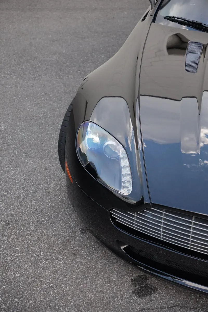 Close-up of a black car's hood and headlight, the paint reflecting the sky and surrounding trees.