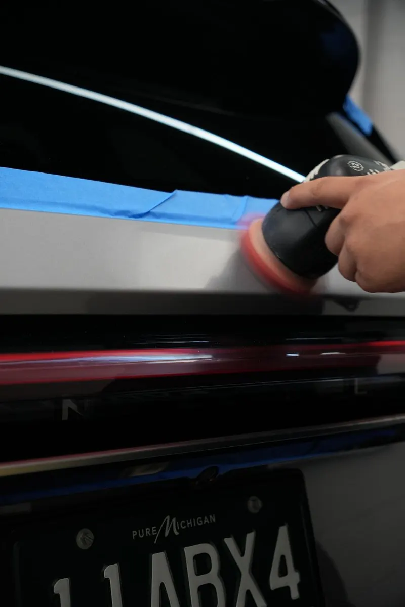 A hand holds a small buffing tool against a vehicle panel near the license plate frame.