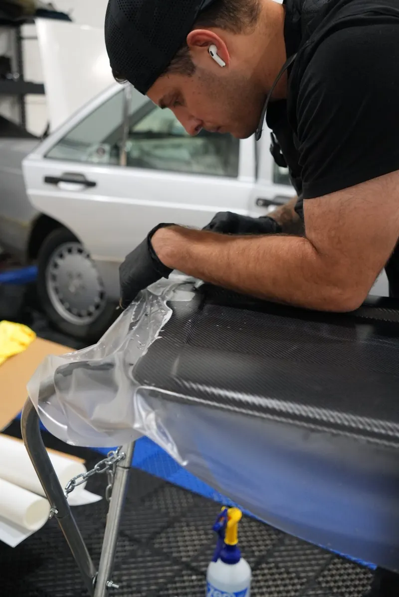 A technician in gloves squeegees a sheet of clear film with slip solution onto a carbon-fiber panel, a spray bottle below and a car parked behind.