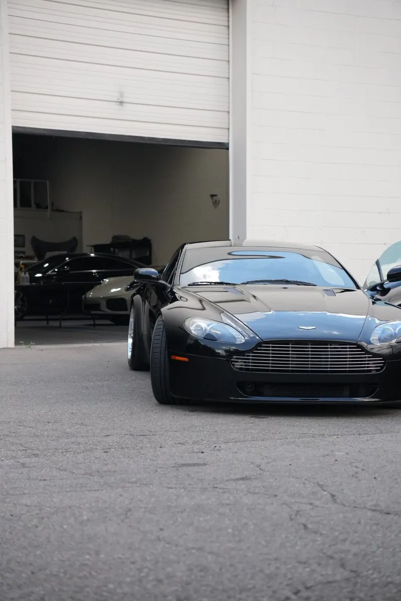 A black car's front end outside an open garage bay, with two more vehicles parked inside.