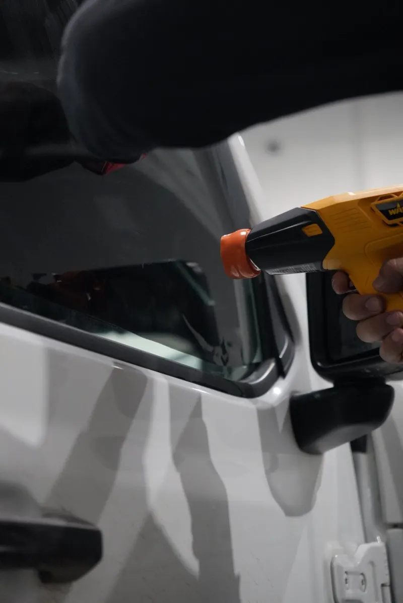 A technician aims a heat gun at the rear quarter window of a white vehicle where film has been applied.