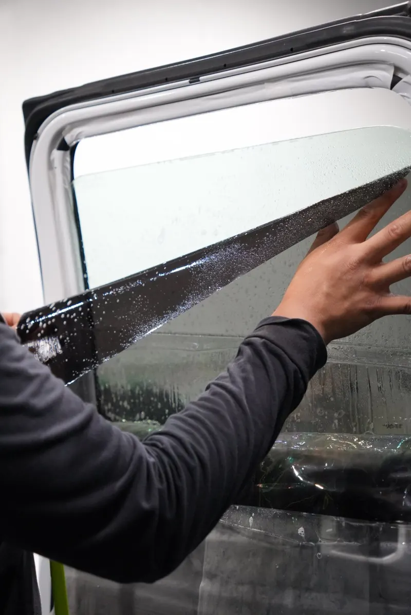 A hand presses a strip of dark film onto the upper corner of a vehicle's wet window.