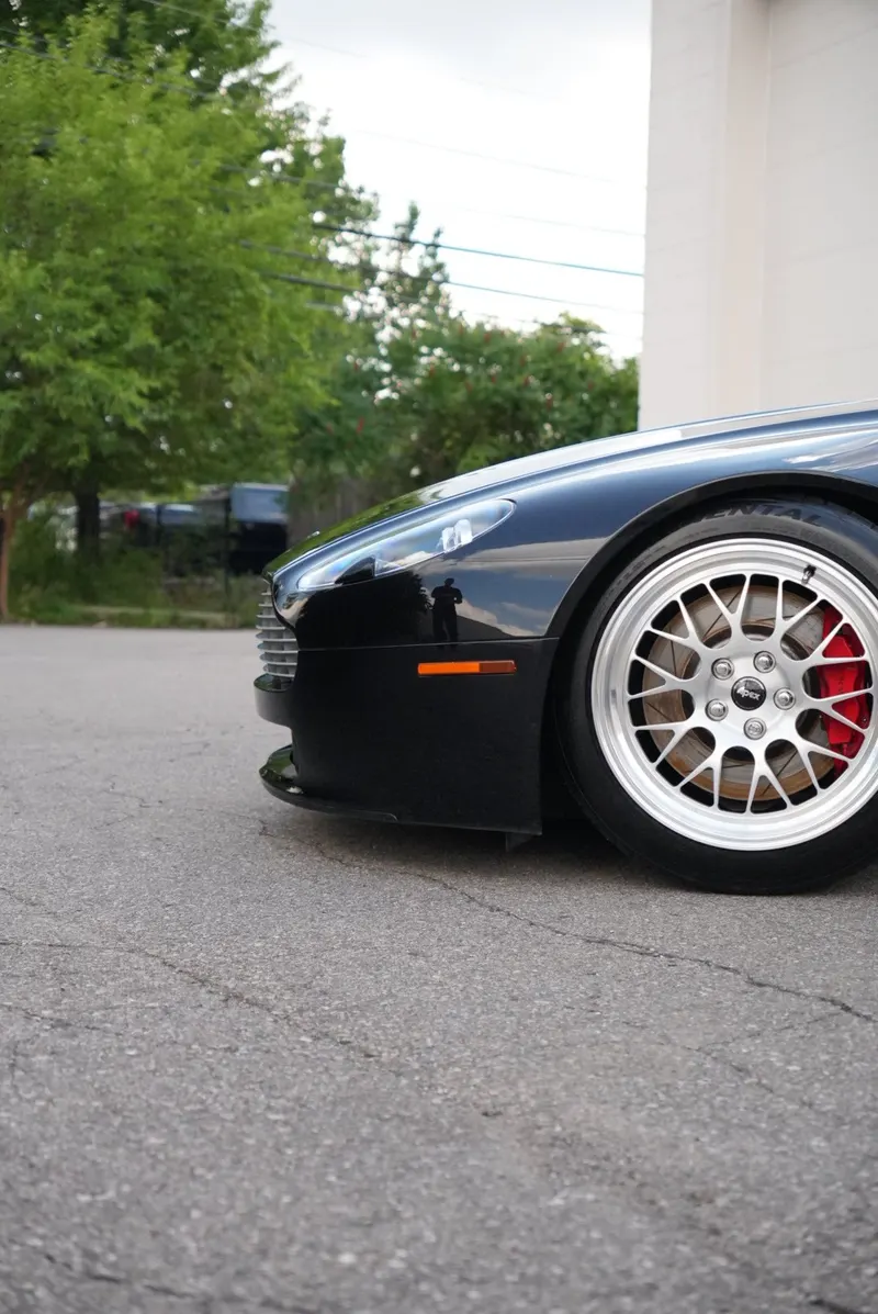 A low-angle close-up of a car's front wheel with a silver multi-spoke rim, red brake caliper, and tire, a building and trees behind.